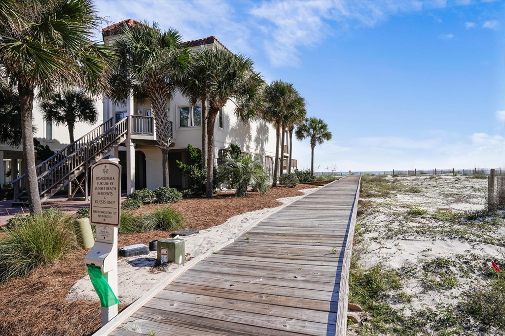 Coastal house with palm trees beside a wooden boardwalk through sandy dunes.