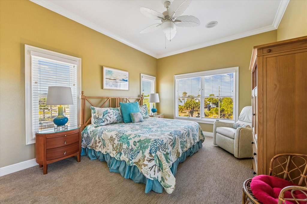 Bedroom with a tropical-patterned bedspread, mustard-yellow walls, and windows overlooking greenery.