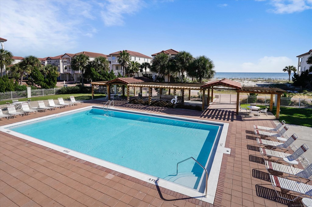Community pool with rows of lounge chairs and a pergola, with the ocean beyond.
