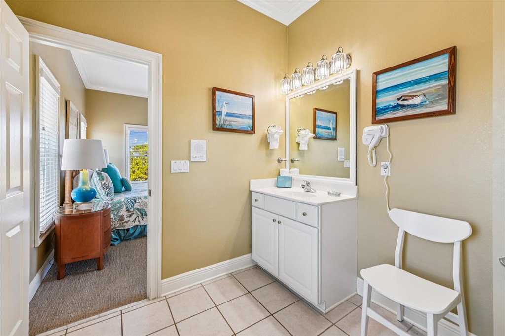 Bathroom with a white vanity and mirror, beach artwork, and an open doorway to a bedroom.