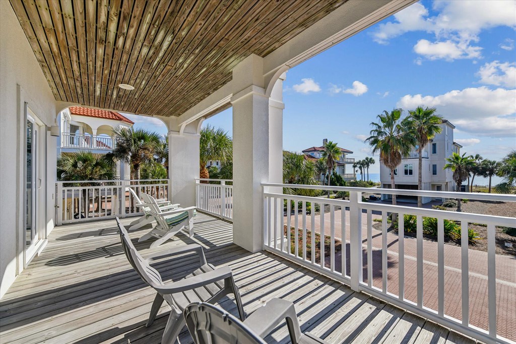 Covered balcony with lounge chairs, white railings, and palms and neighboring homes beyond.