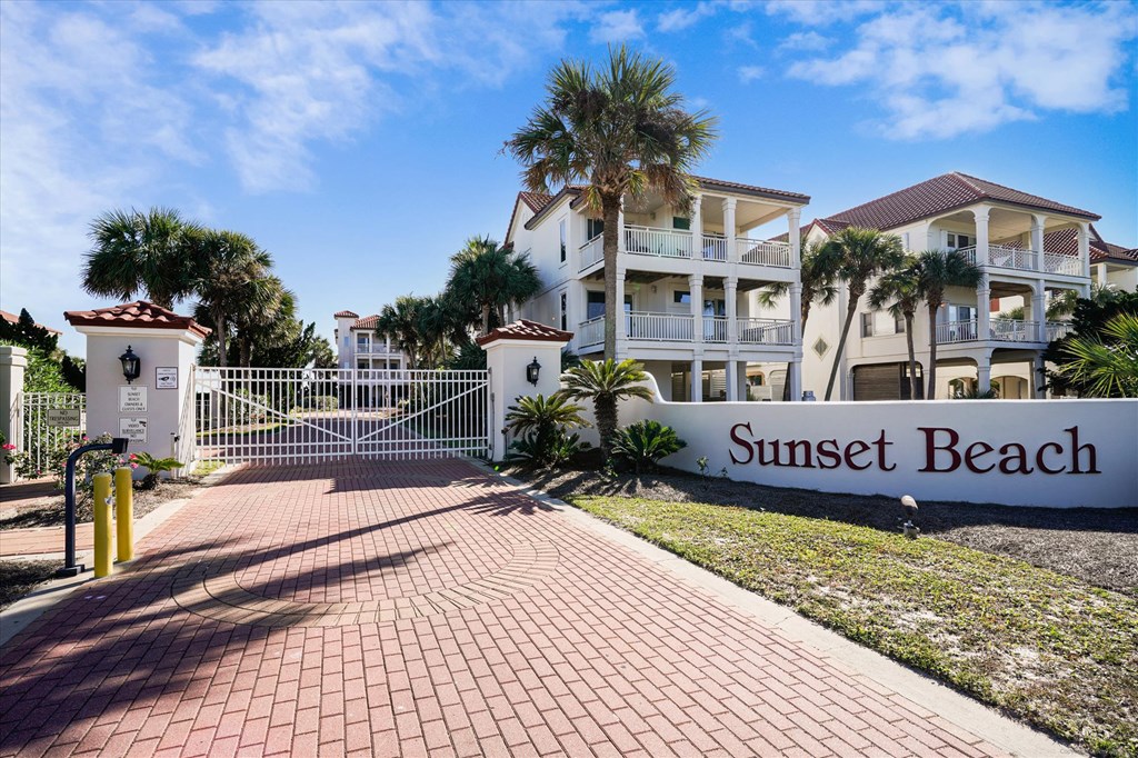 Gated Sunset Beach entrance with a brick drive, palm trees, and coastal homes.
