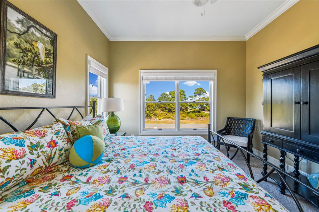 Bedroom with a colorful floral quilt, dark armoire, and window overlooking coastal vegetation.