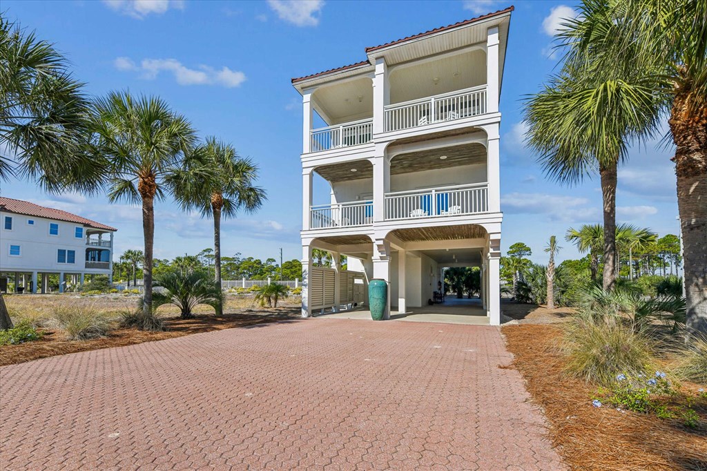 Elevated coastal home with stacked balconies, white railings, palm trees, and a brick-paver driveway.