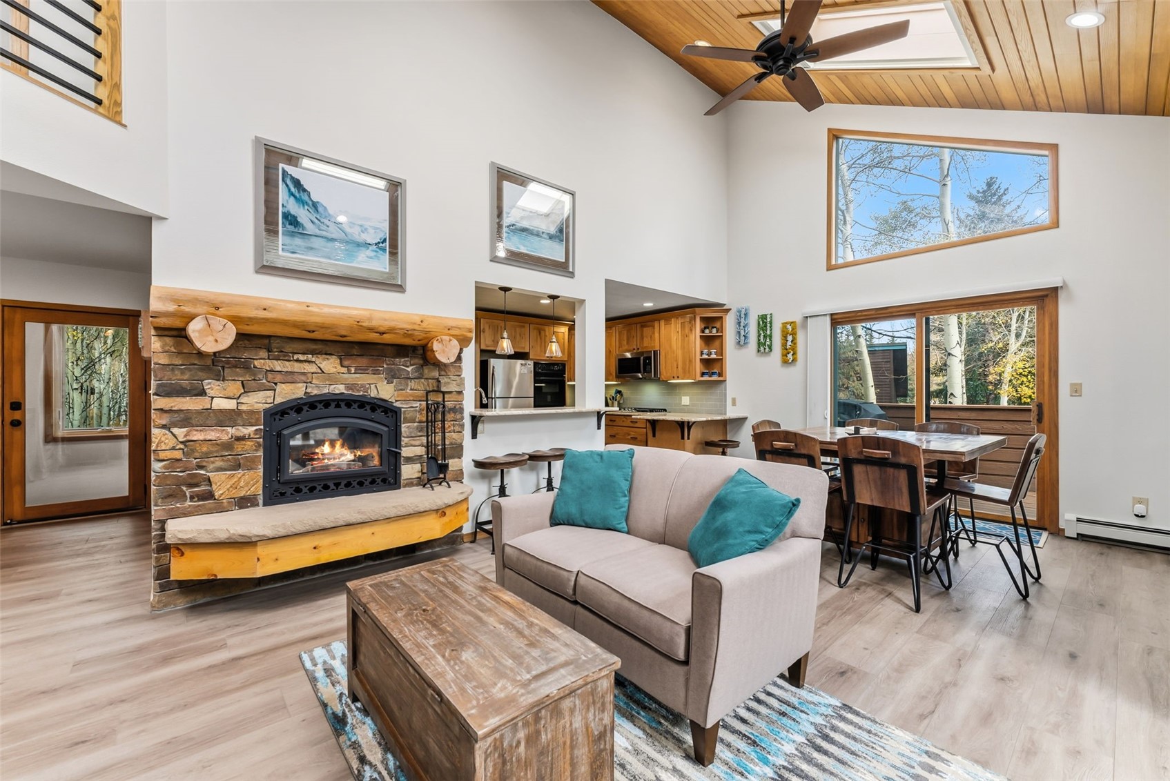 Open living and dining area with a stone fireplace, vaulted wood ceiling, and large windows.