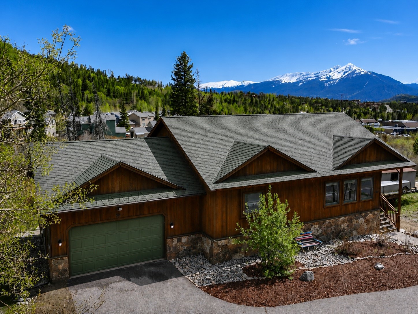 Wood-sided home with a green garage door and gabled roof, backed by snow-capped mountains.