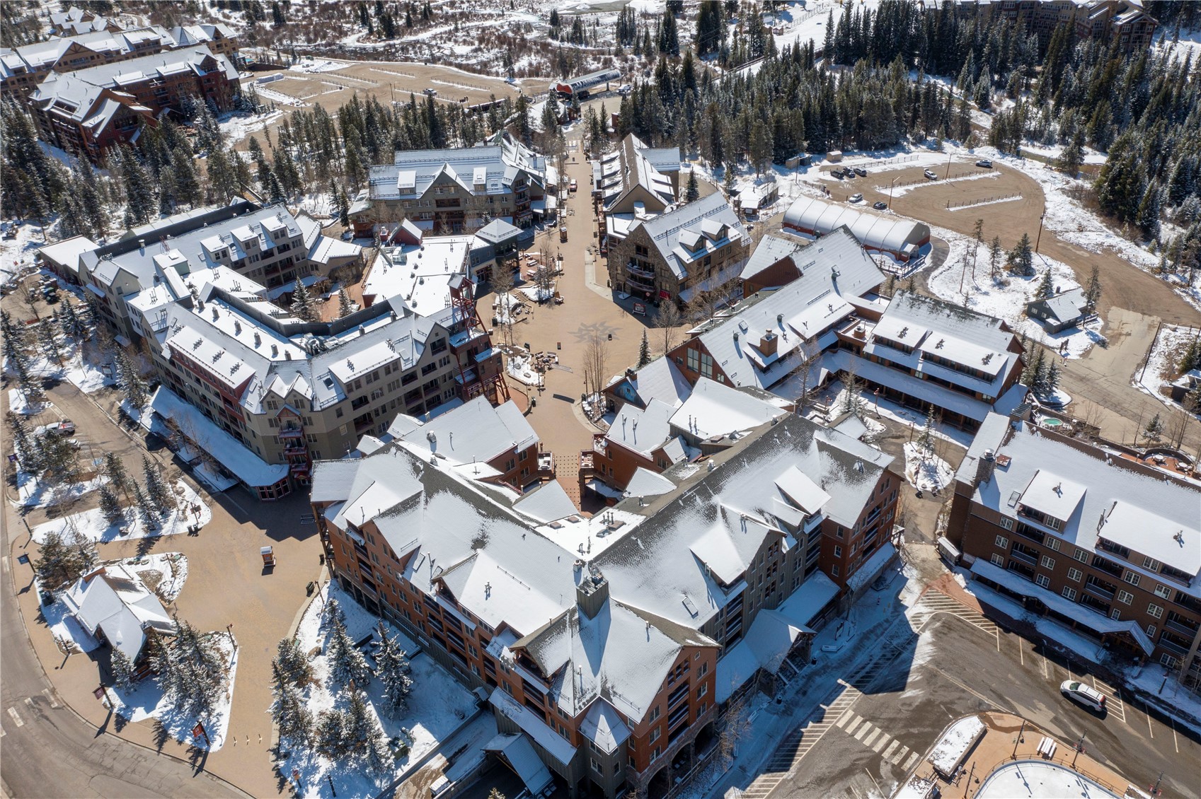 Aerial view of snow-covered resort buildings around a central plaza, surrounded by evergreen trees.