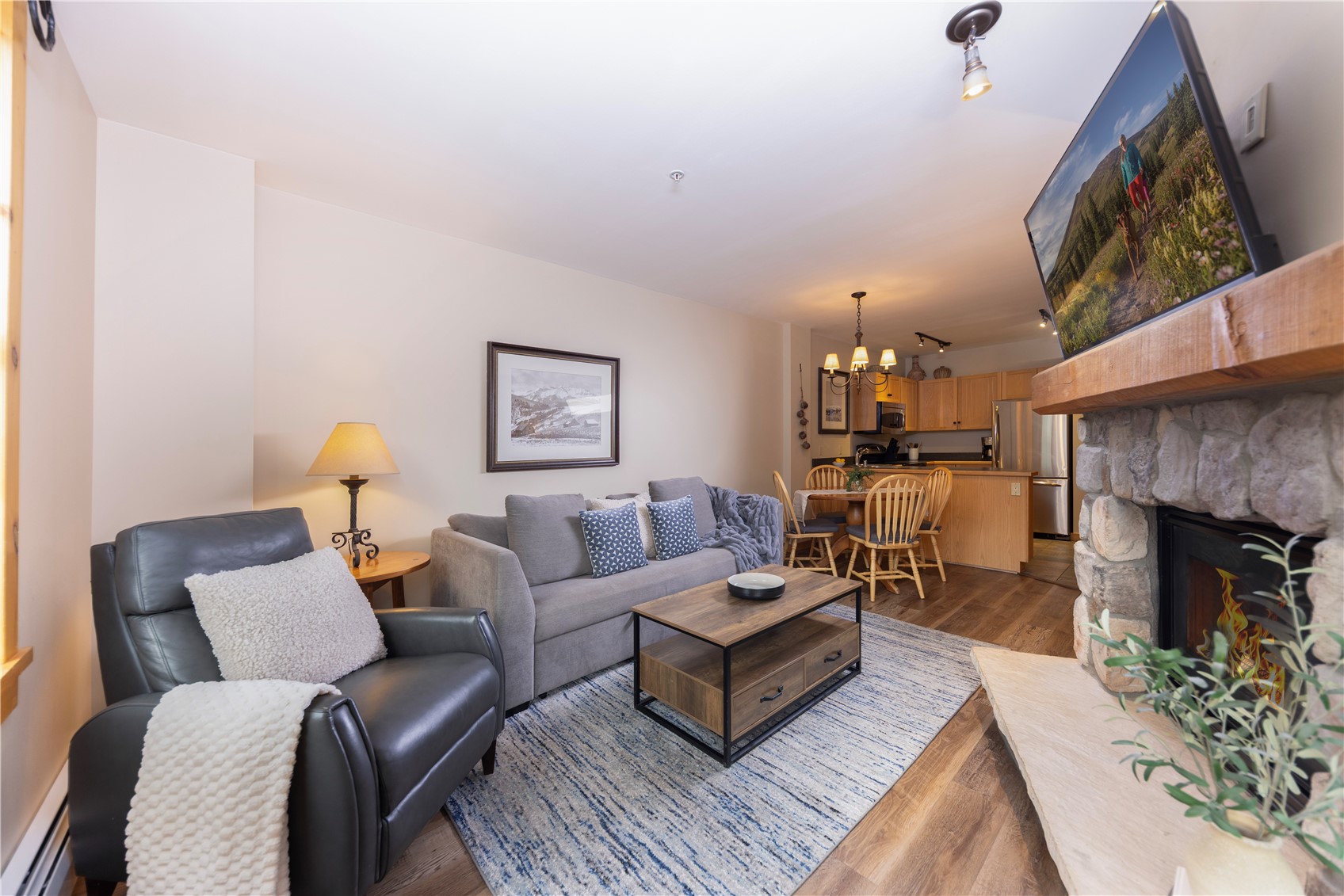 Furnished living room with a stone fireplace, gray sofa, and open kitchen and dining area.