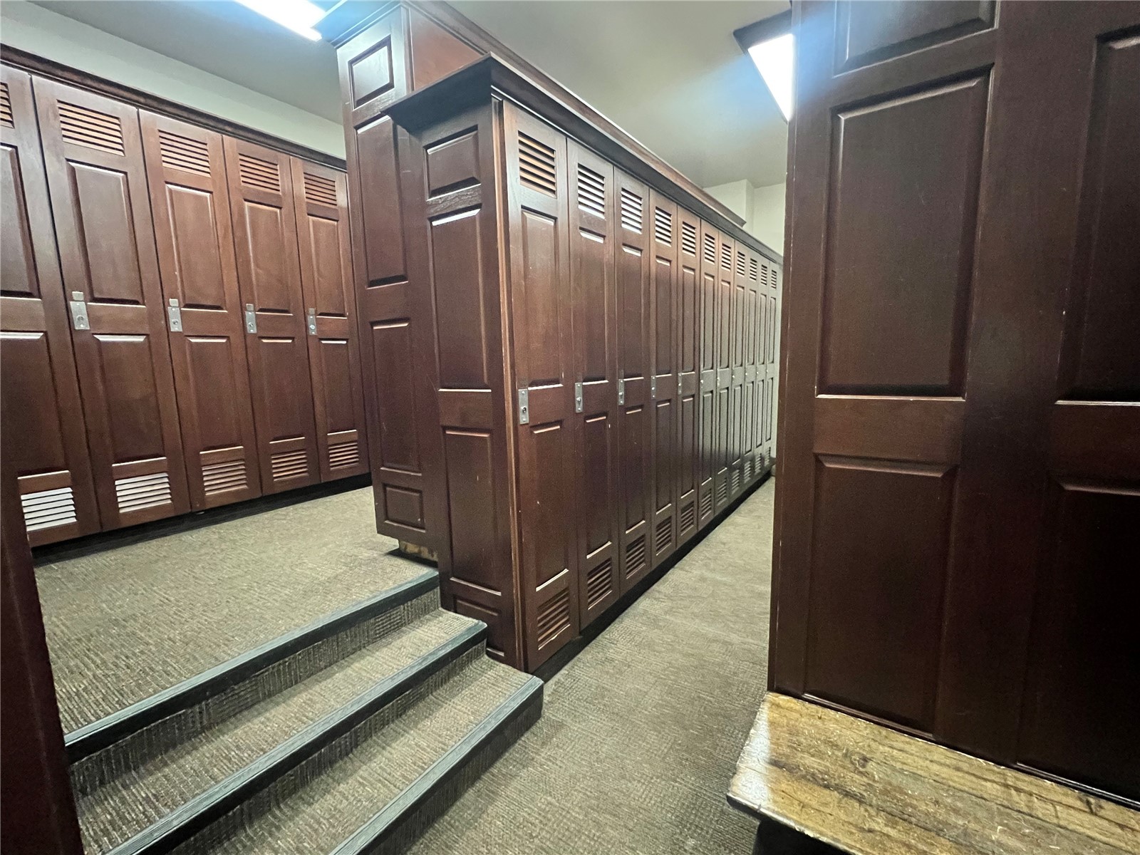 Locker room with rows of dark wooden lockers, carpeted steps, and a wooden bench.