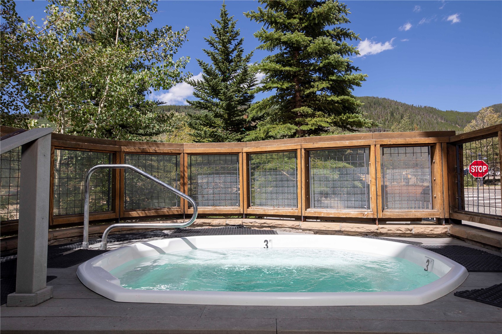 Outdoor hot tub on a deck, with a wooden fence and forested mountain slopes beyond.