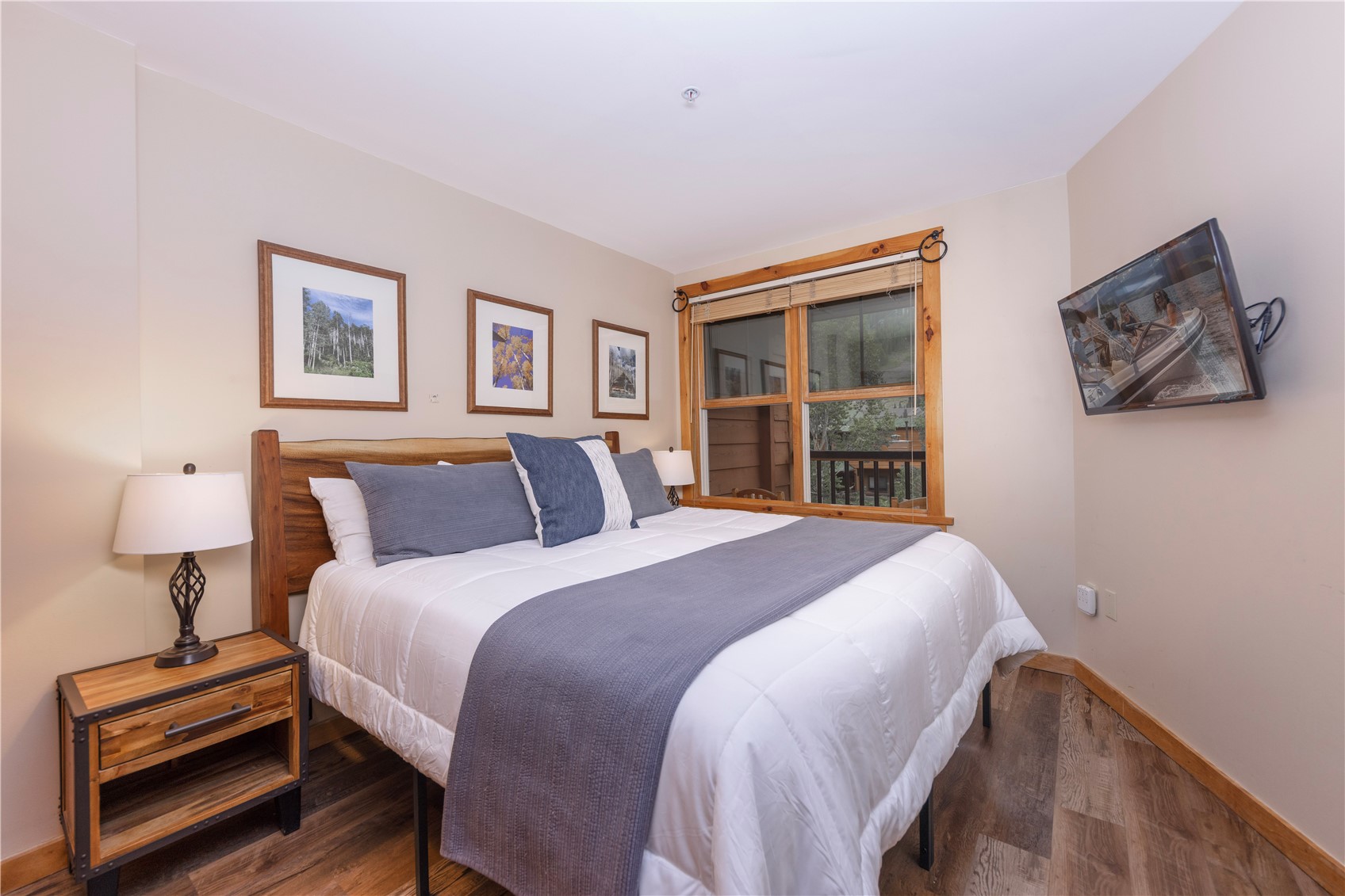 Bedroom with a wooden bed, gray-and-white bedding, framed artwork, and a window overlooking trees.