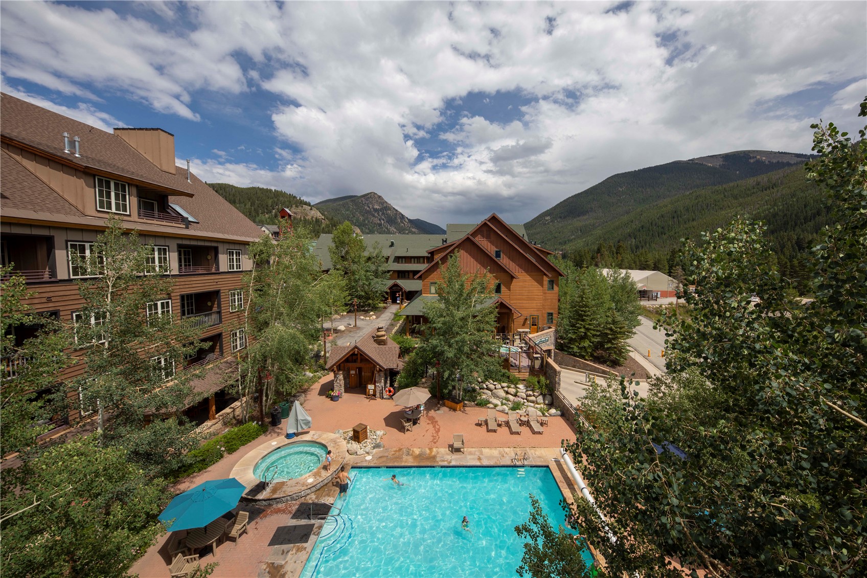 Outdoor pool and round hot tub among lodge buildings, with forested mountains in the background.