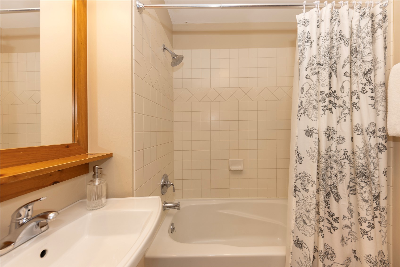 Bathroom with a tiled tub and shower, floral curtain, and wooden-framed mirror.