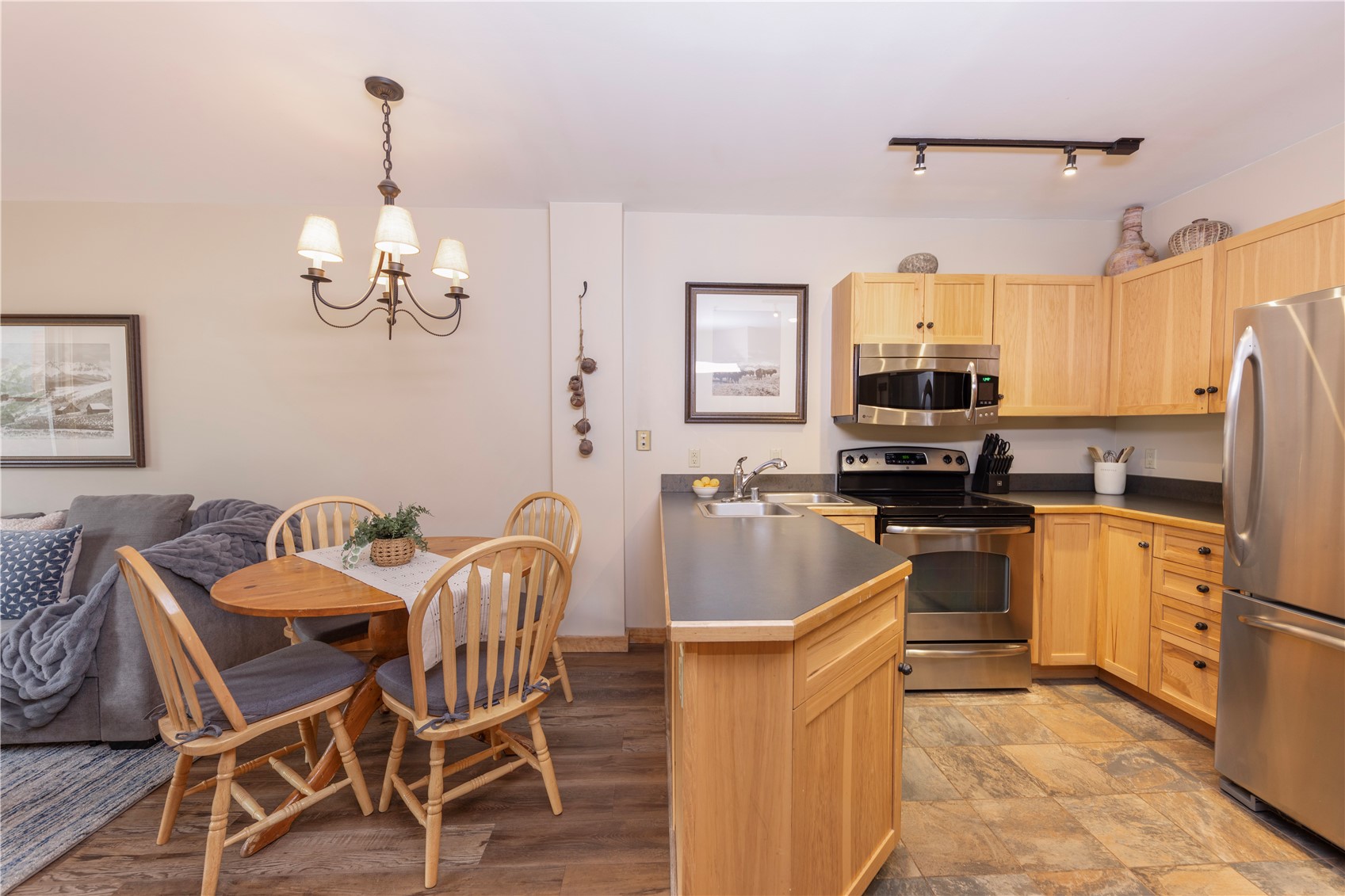 Open-plan kitchen with light wood cabinets, stainless steel appliances, and a dining table beside a gray sofa.