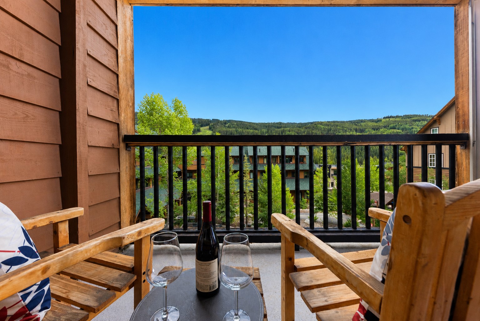Furnished balcony overlooking forested hills, with wooden chairs and wine glasses on a small table.
