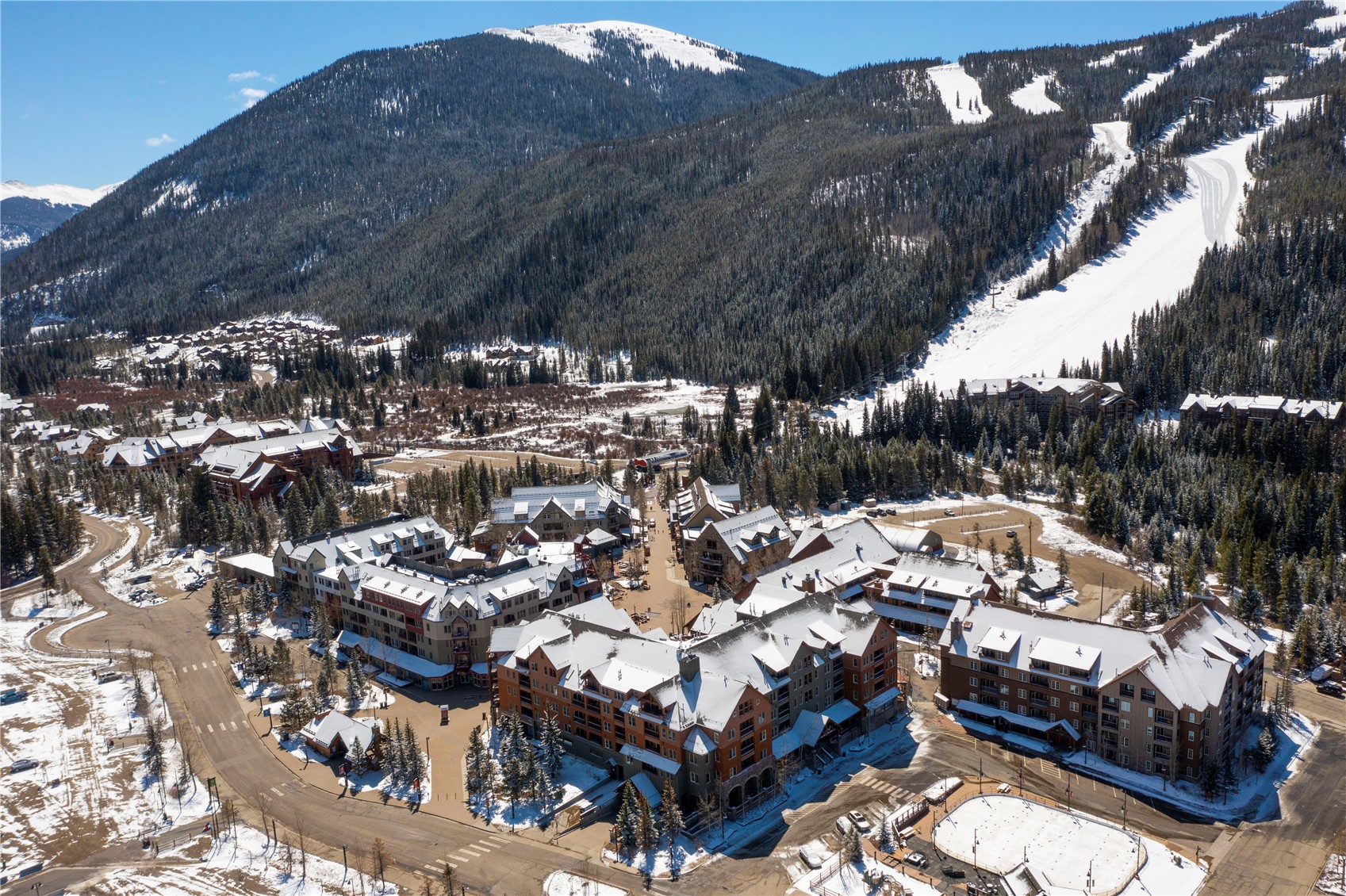 Mountain resort village below forested ski slopes, with snow-covered buildings and groomed runs.