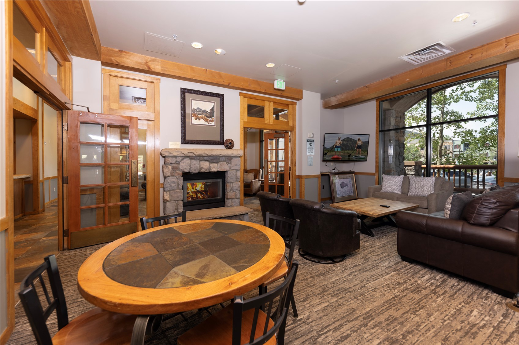 Furnished living room with a stone fireplace, round dining table, and large windows overlooking a deck.