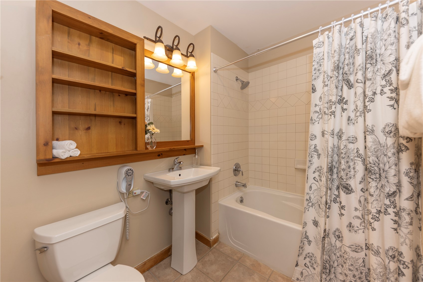 Bathroom with a white tub, floral shower curtain, pedestal sink, and built-in wooden shelving.