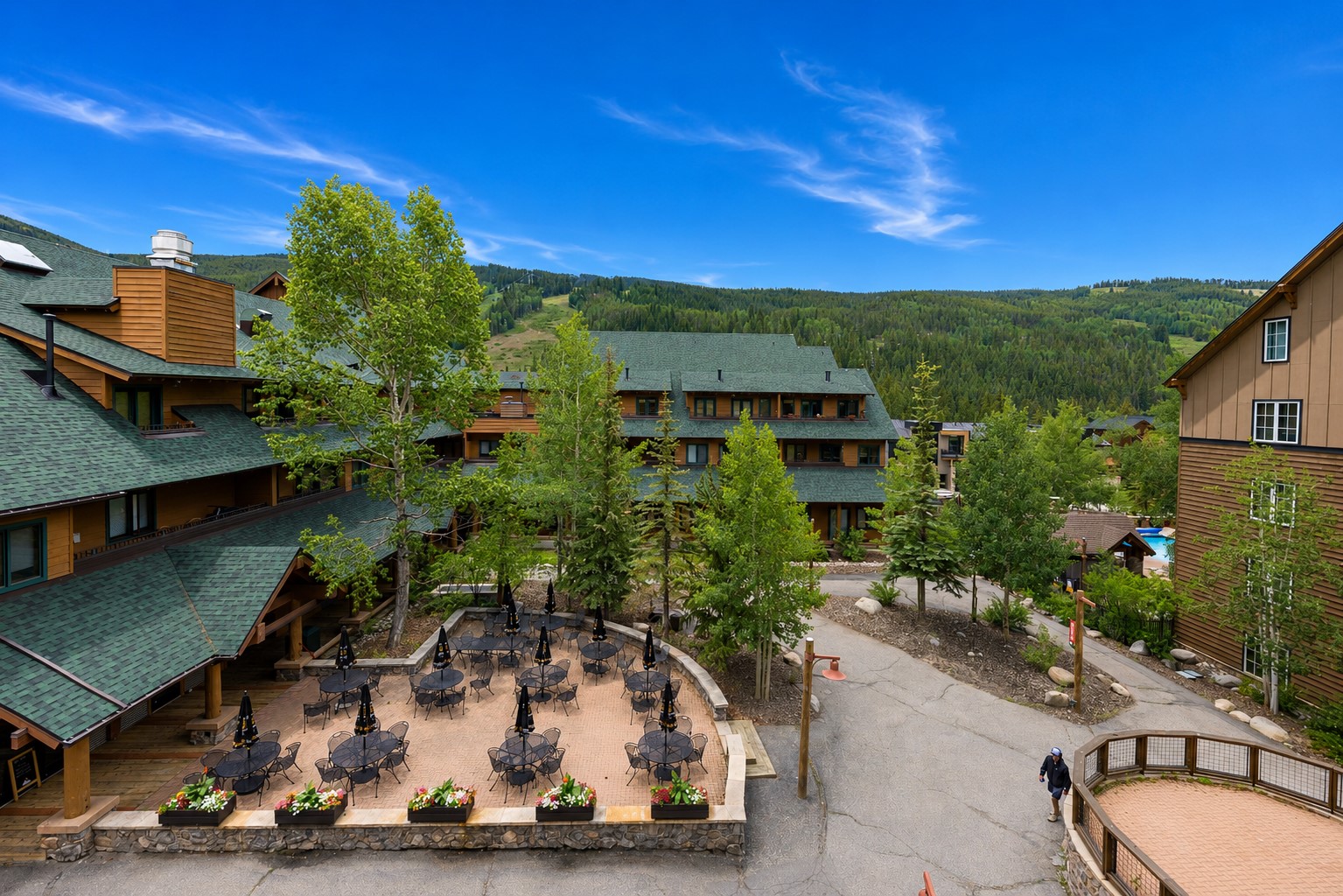 Mountain resort buildings surround a courtyard with outdoor dining tables and forested hills in the background.