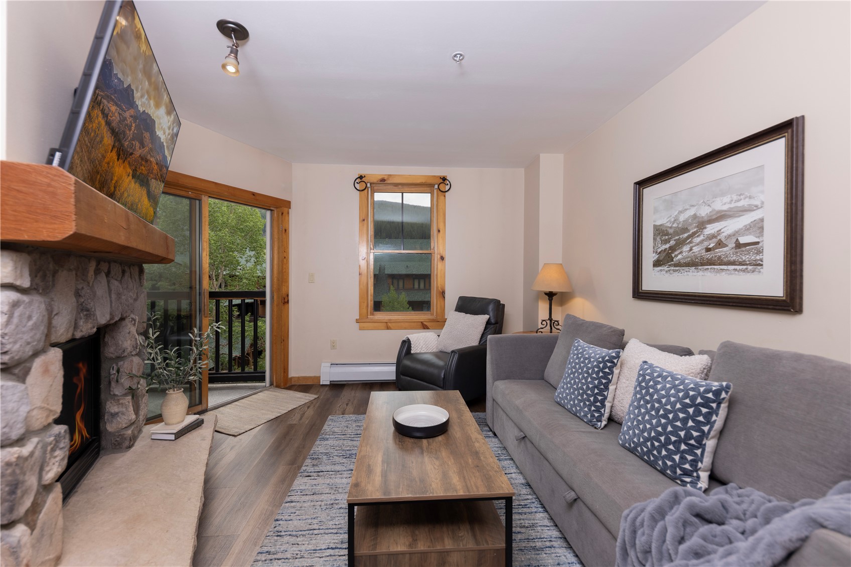 Living room with a stone fireplace, gray sofa, and sliding glass door overlooking trees.