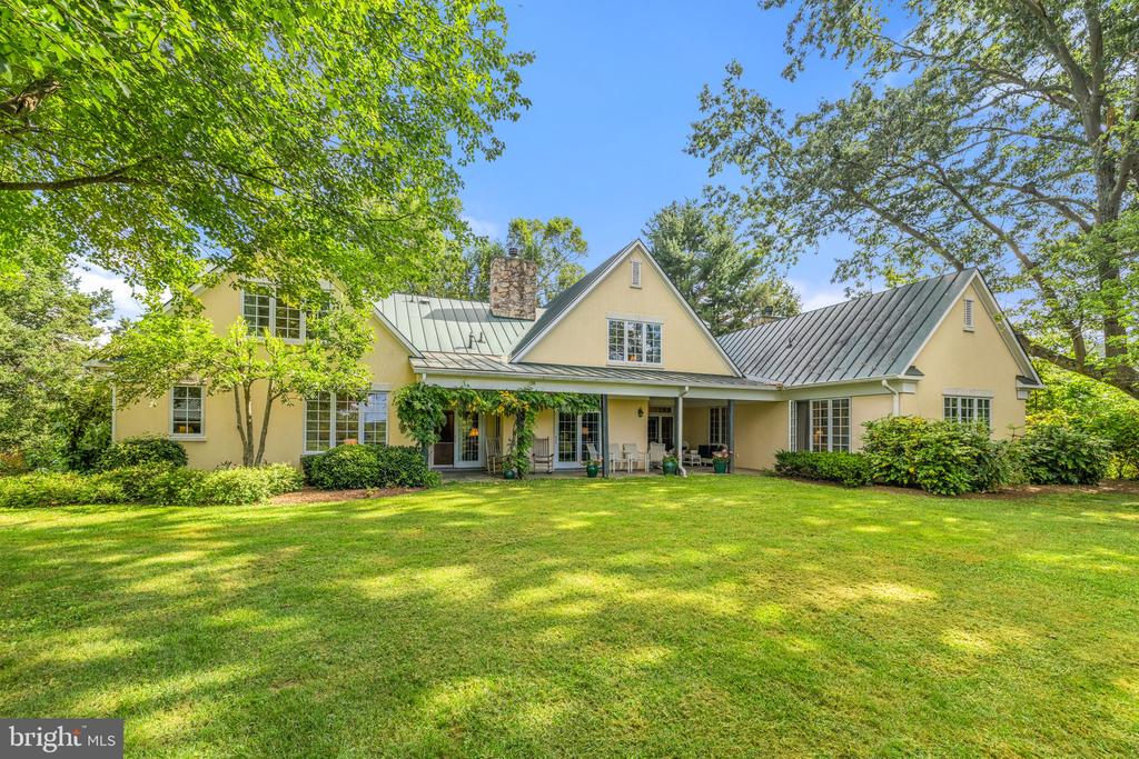 Pale yellow country home with a metal roof, stone chimney, covered patio, and broad lawn.
