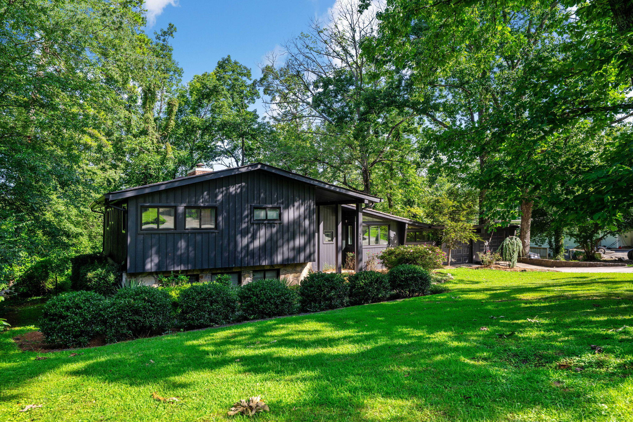 Dark, low-profile home with broad windows and a covered entry among mature trees and a sloping lawn.