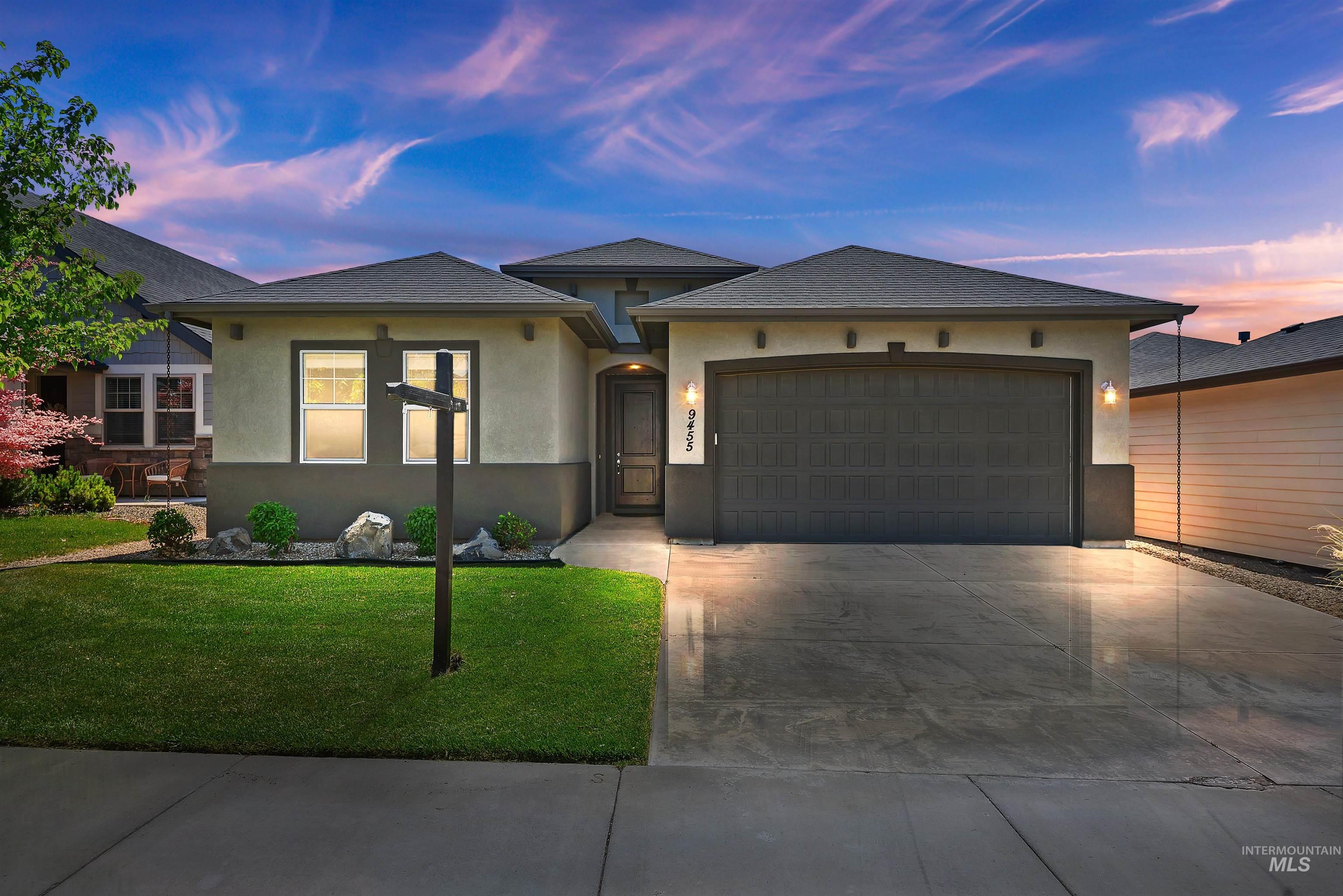 Single-story stucco home with dark double garage, broad driveway, and green front lawn at dusk.