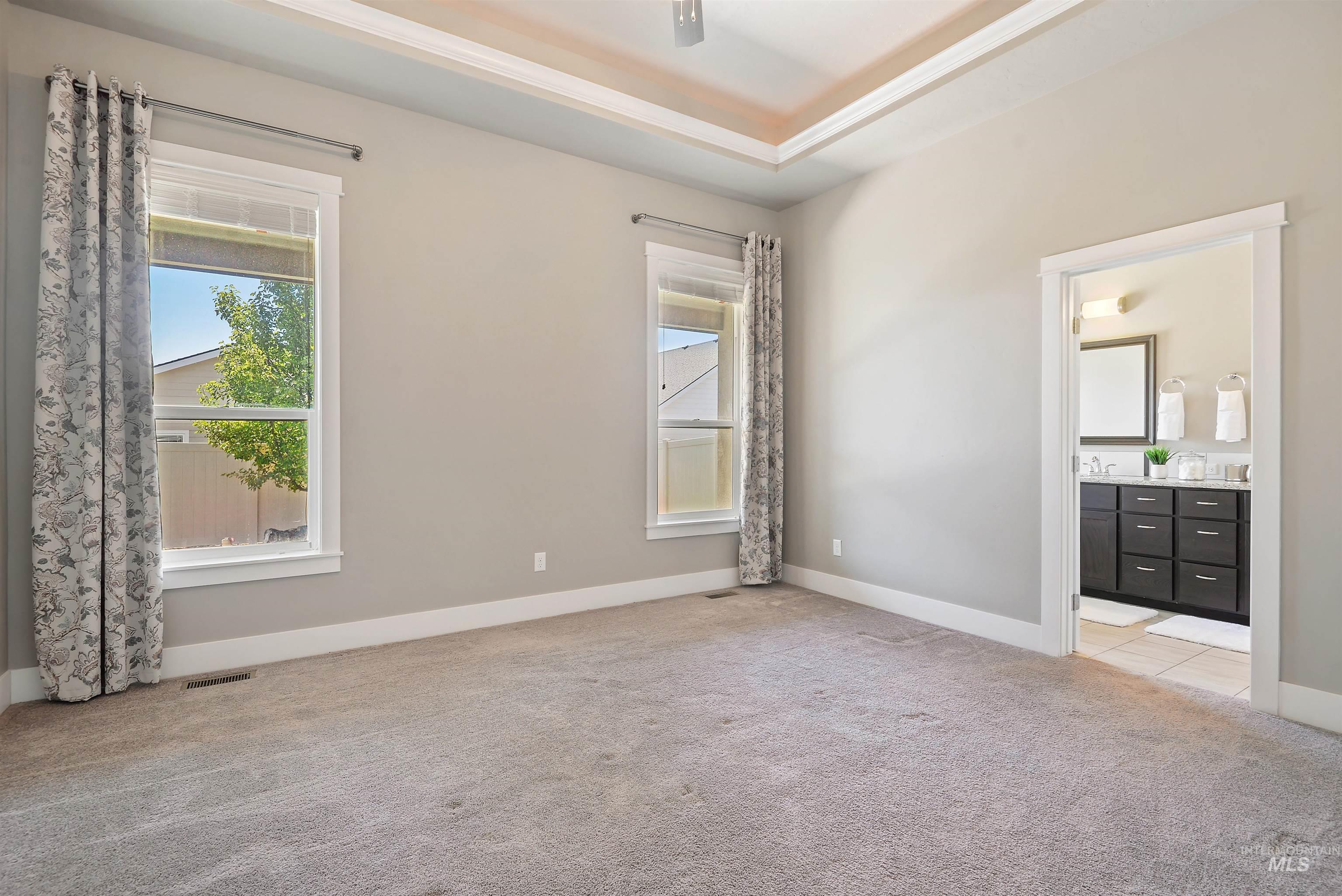 Empty bedroom with a tray ceiling, patterned curtains, and an open doorway to a bathroom.