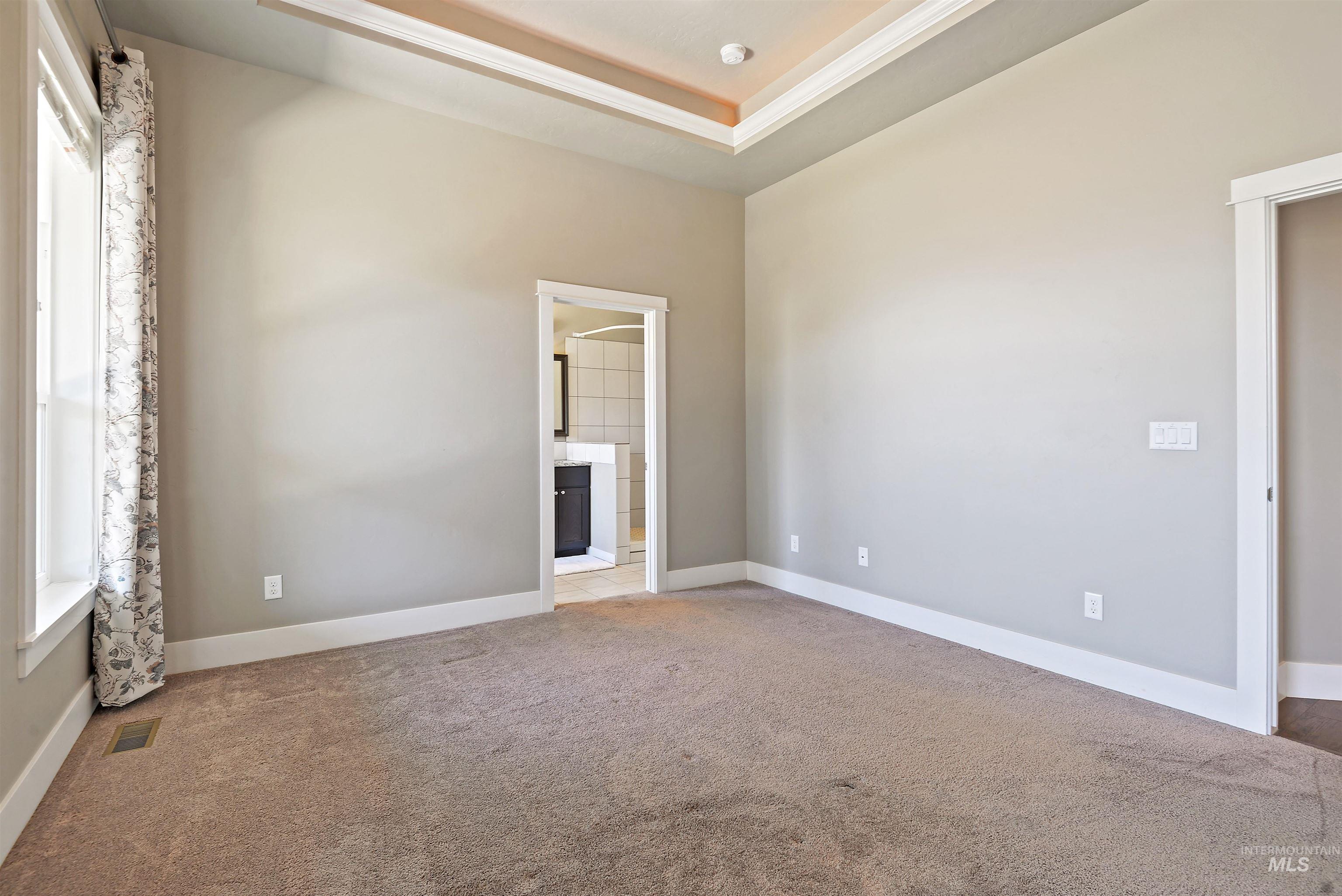 Empty bedroom with a tray ceiling, patterned curtains, and an ensuite bathroom doorway.