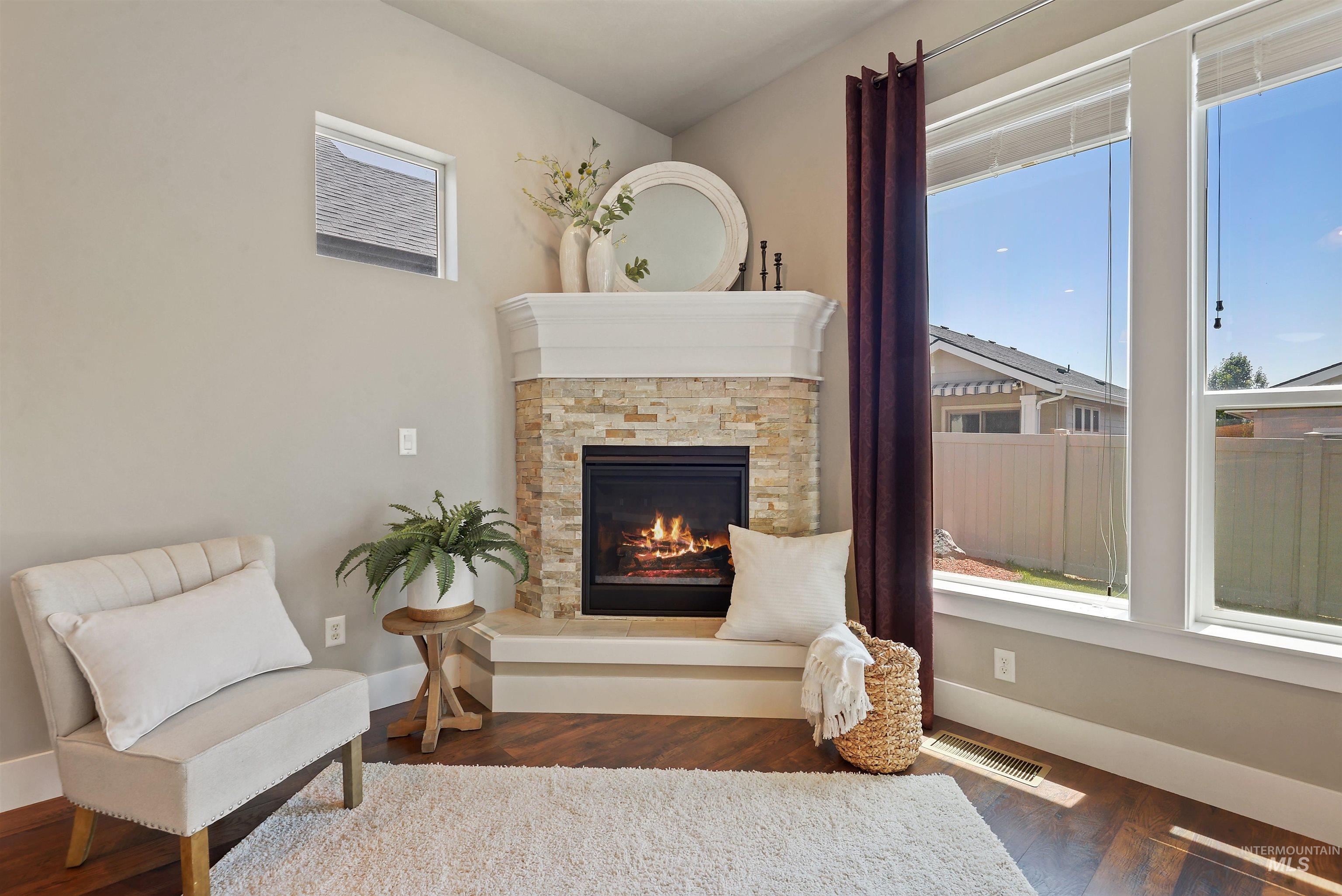 Staged living room with a stone corner fireplace, lit fire, and large windows overlooking a fenced yard.