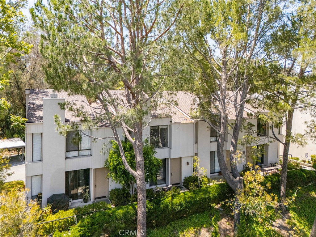 Two-story light-colored residence screened by tall trees, with landscaped grounds and patio seating.