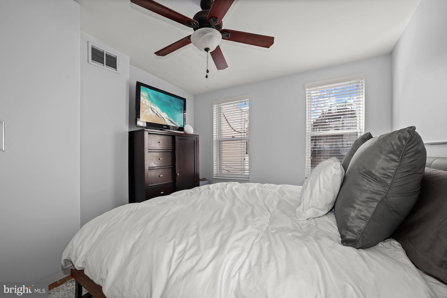 Bedroom with white bedding, gray pillows, a dark wood dresser, and two windows with blinds.