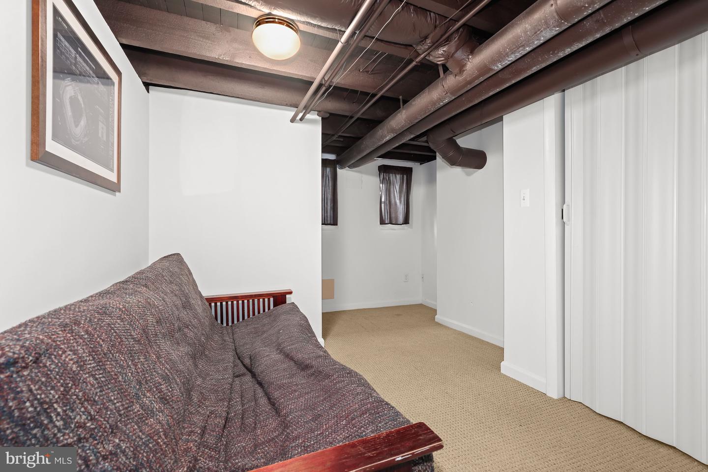 Basement sitting area with a patterned sofa, beige carpet, and exposed ceiling ducts.