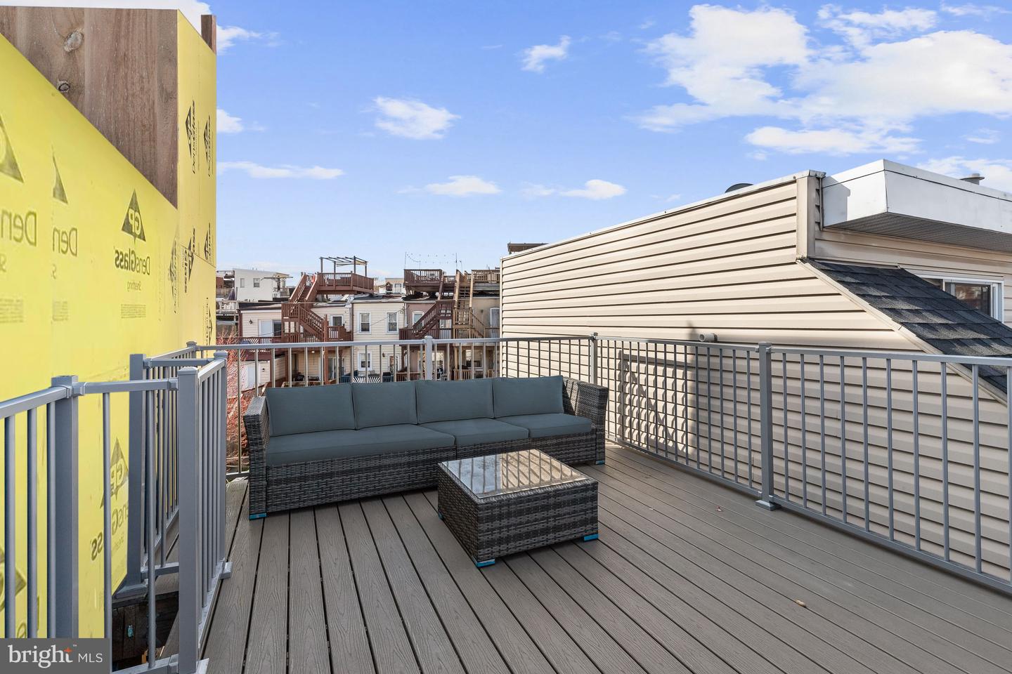 Rooftop deck with wicker seating, gray railings, and views of neighboring rowhouse rooftops.