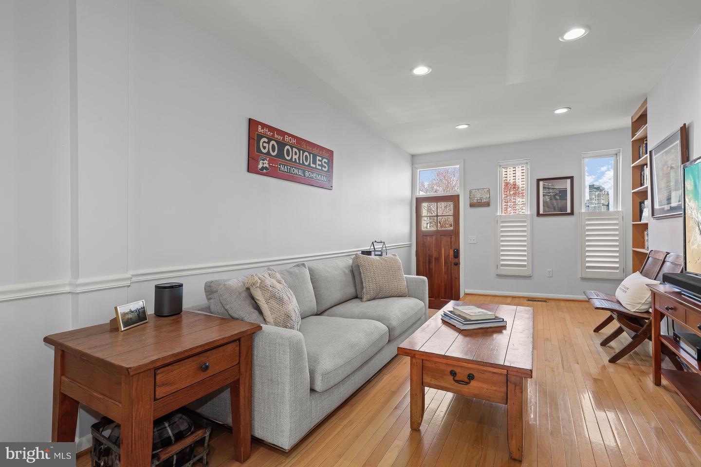 Living room with a gray sofa, warm hardwood floors, and tall windows overlooking nearby buildings.