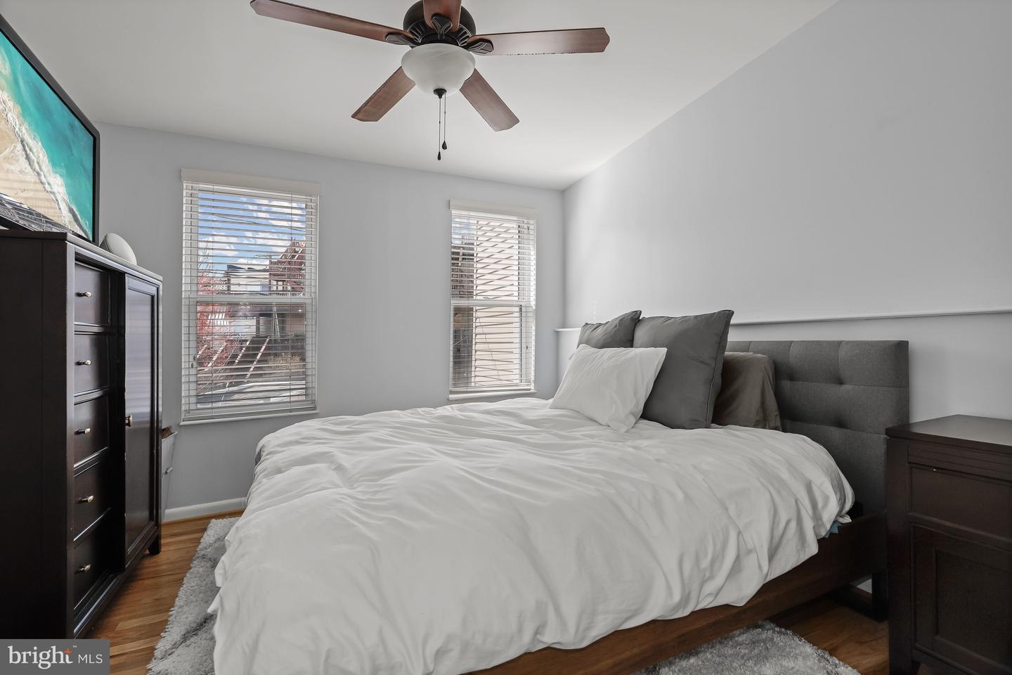 Furnished bedroom with a white bed, gray headboard, hardwood floors, and two windows with blinds.