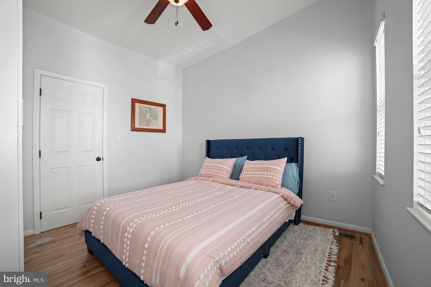 Bedroom with pink striped bedding, a navy tufted headboard, hardwood floors, and windows with blinds.