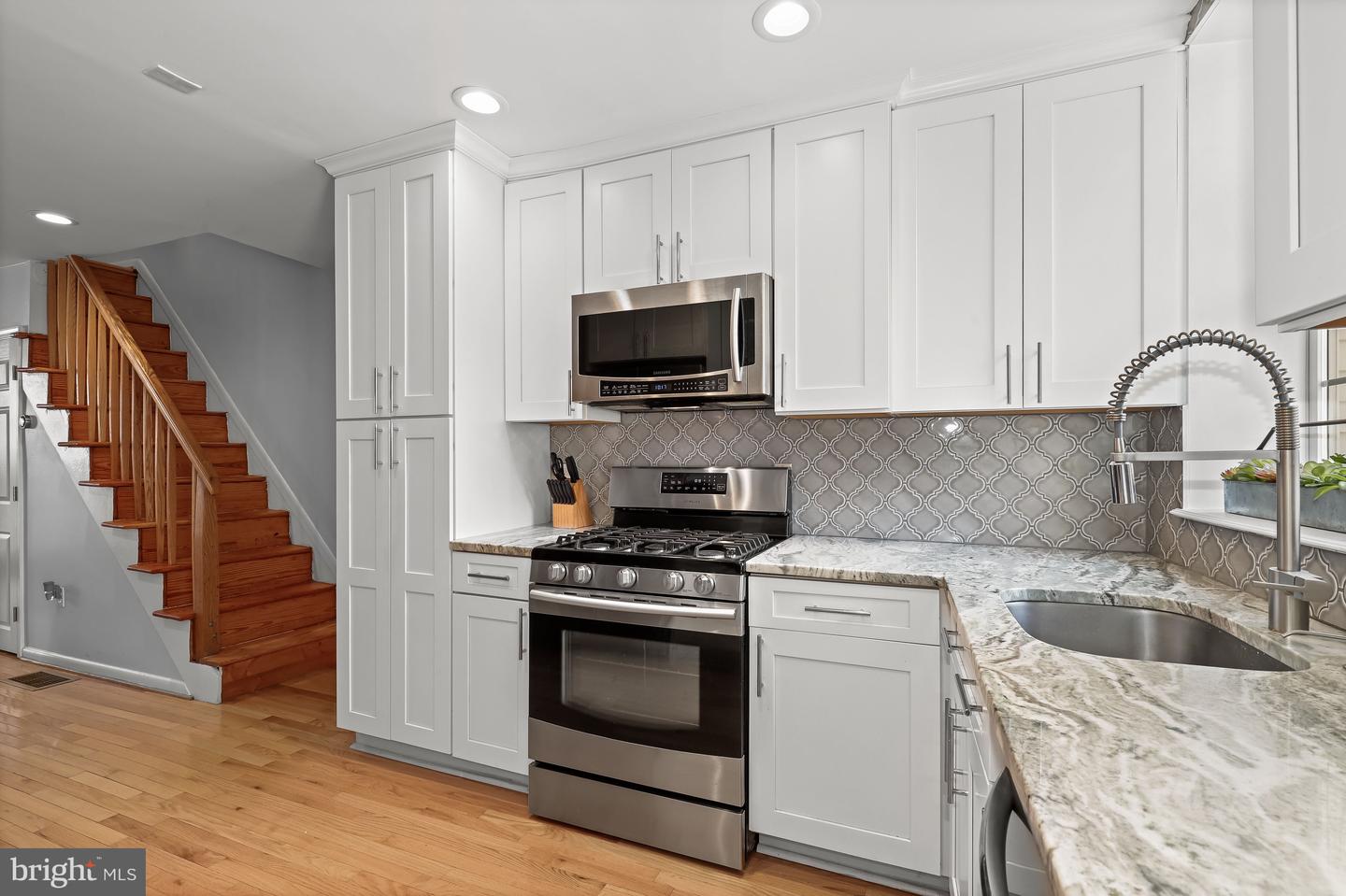 Renovated kitchen with white cabinets, patterned gray backsplash, and hardwood stairs beside the cooking area.