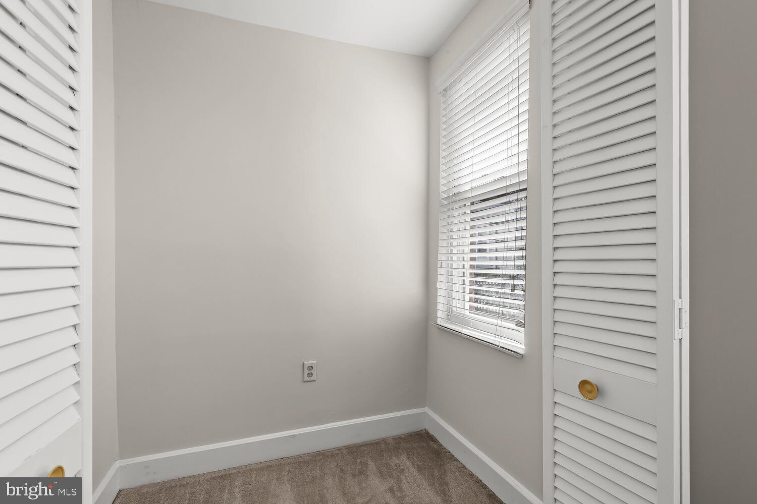 Empty carpeted closet with white louvered doors and a window with blinds.