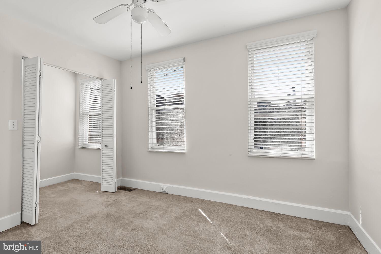 Empty bedroom with beige carpet, two windows with white blinds, and louvered closet doors.