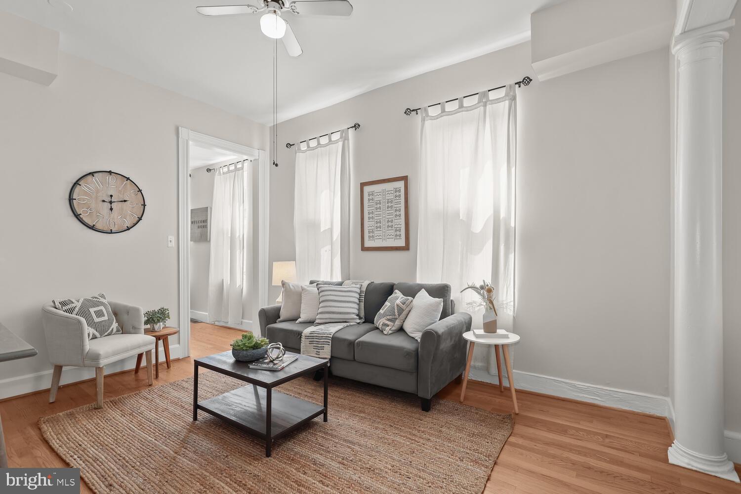 Living room with gray seating, hardwood floors, tall curtained windows, and a ceiling fan.