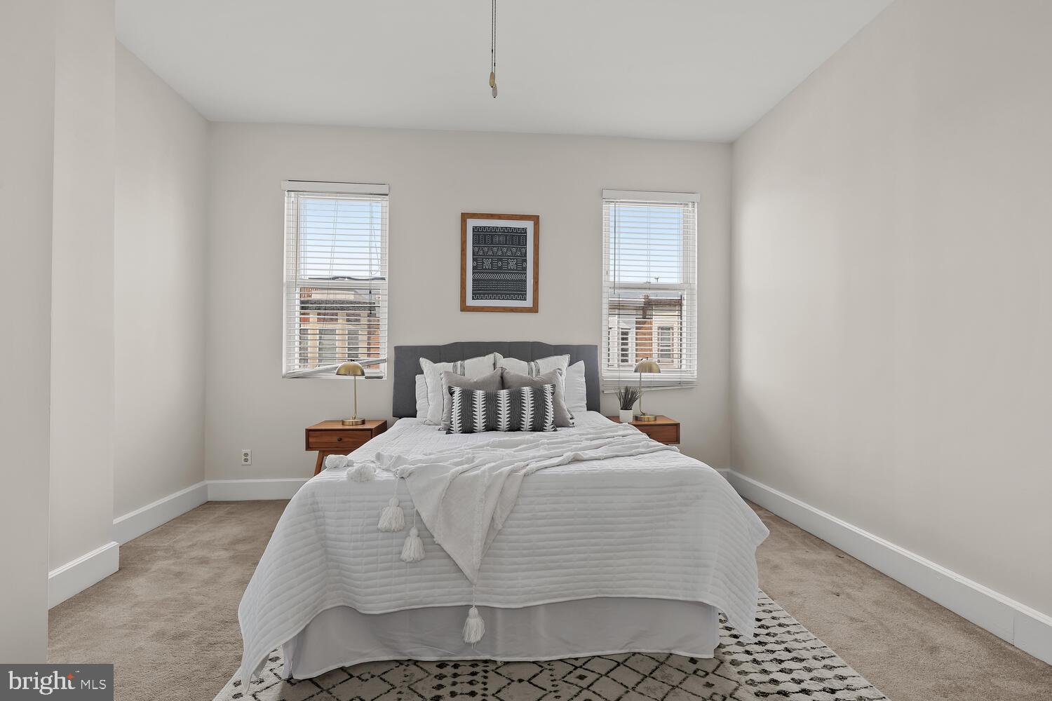 Staged bedroom with a white bed, gray headboard, matching nightstands, and two windows.