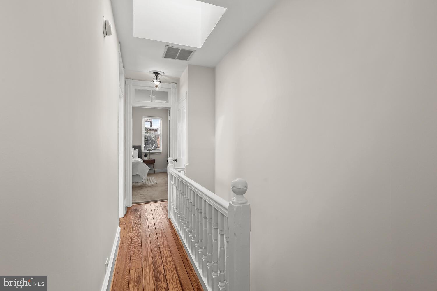Upstairs hallway with polished wood floors and white railing, looking toward a bedroom.