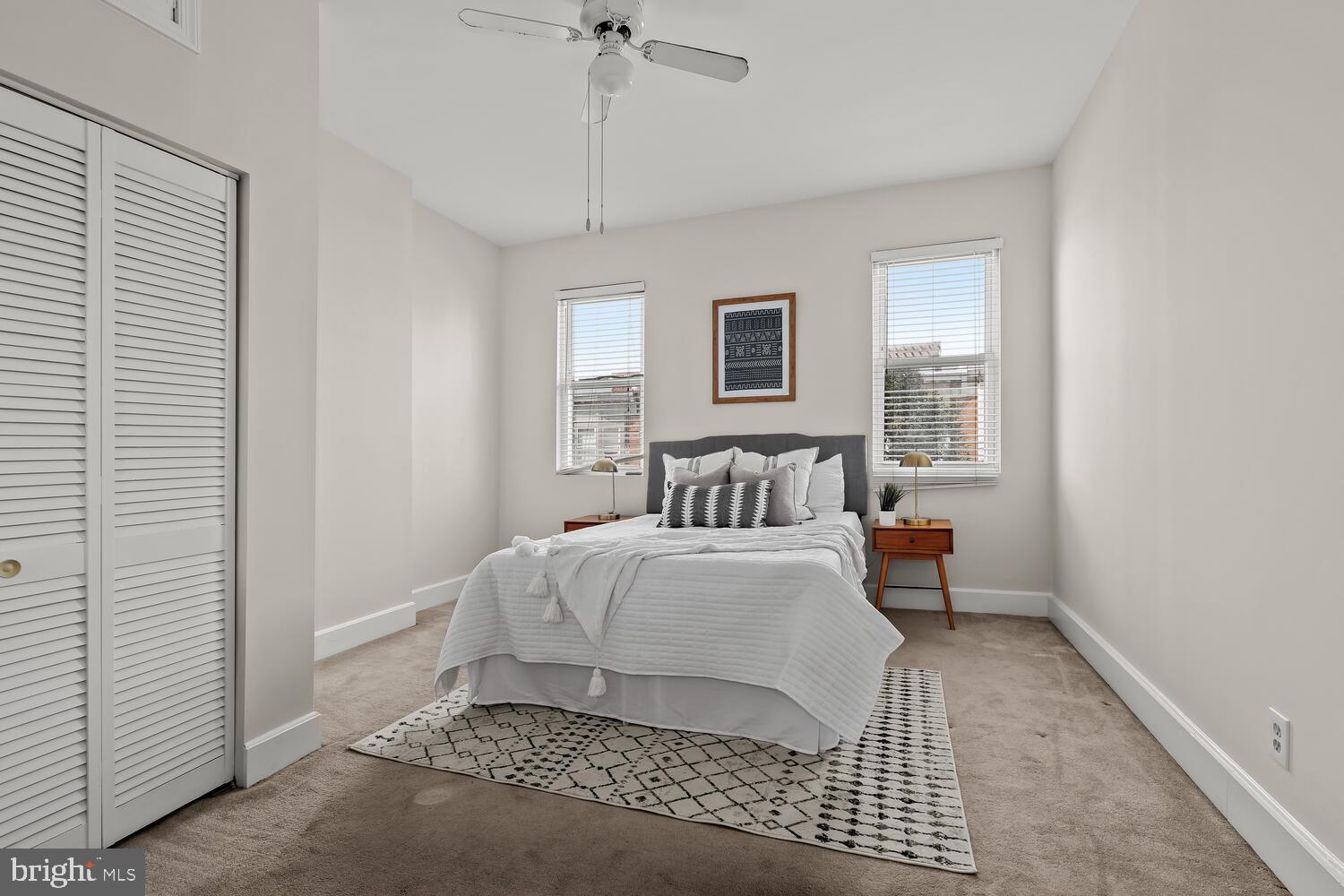 Staged bedroom with white bedding, two windows, wooden nightstands, and a patterned rug.