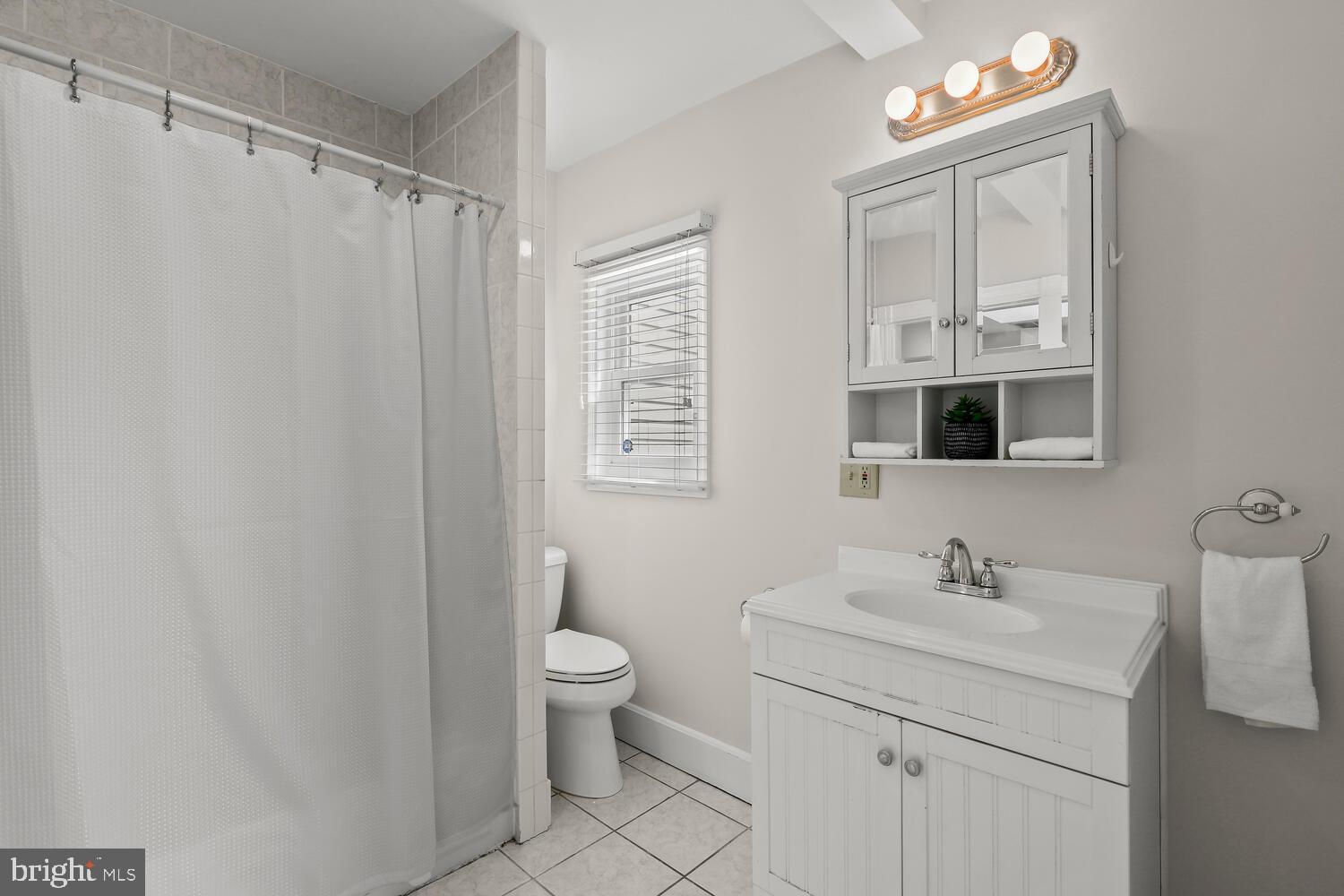Bathroom with white vanity and mirrored cabinet, tiled shower, and window with blinds.