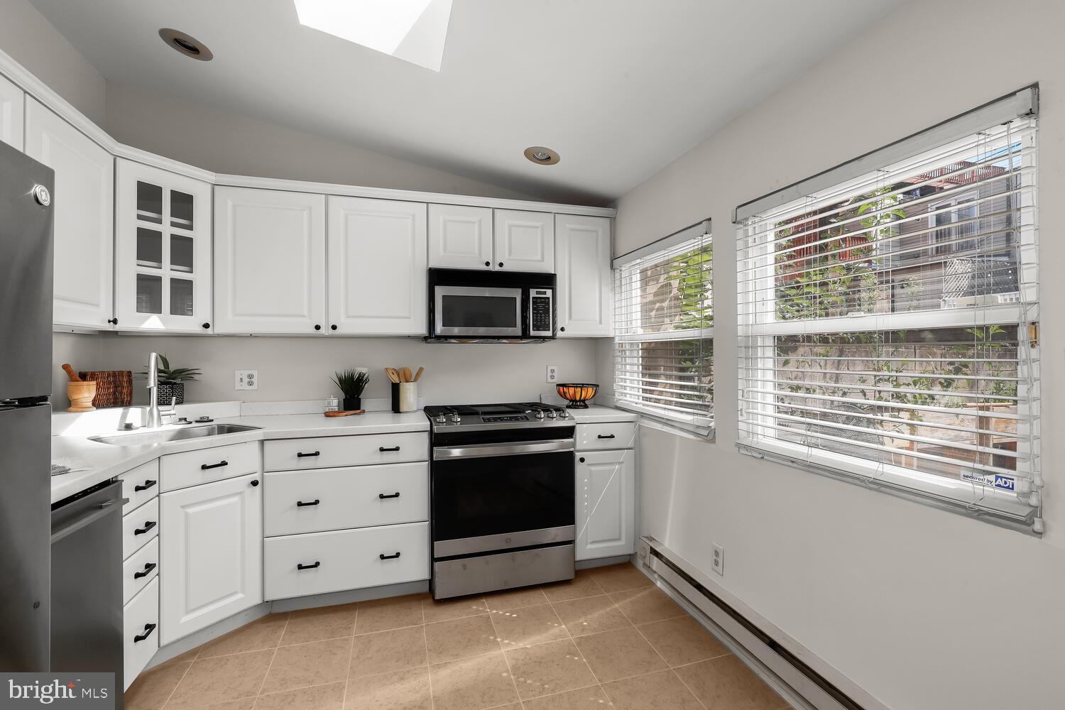 Kitchen with white cabinets, stainless-steel appliances, beige tile flooring, and two large windows.