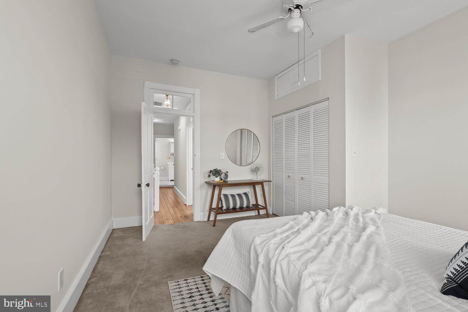 Staged bedroom with a white bed, louvered closet doors, and a console beneath a round mirror.