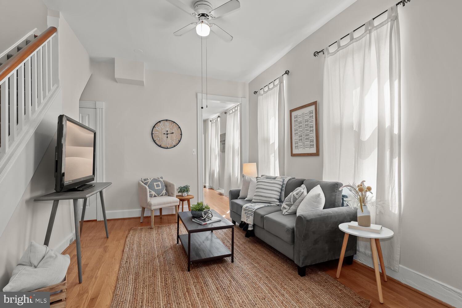Staged living room with a gray sofa, tall curtained windows, and a staircase.
