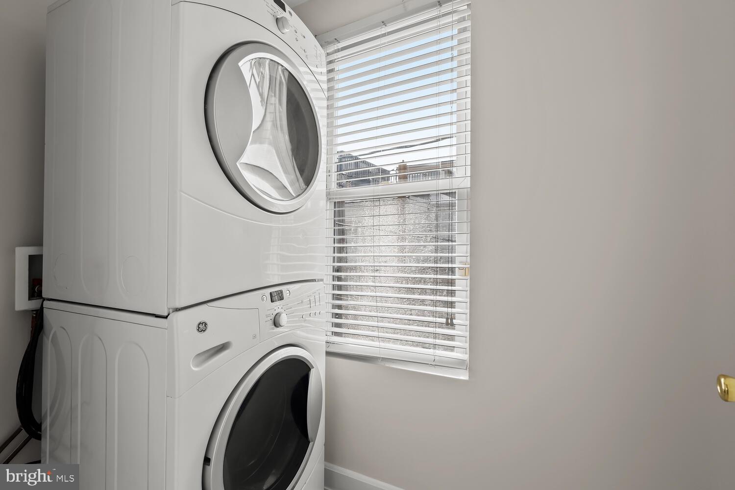 Laundry area with stacked white front-loading washer and dryer beside a window with blinds.