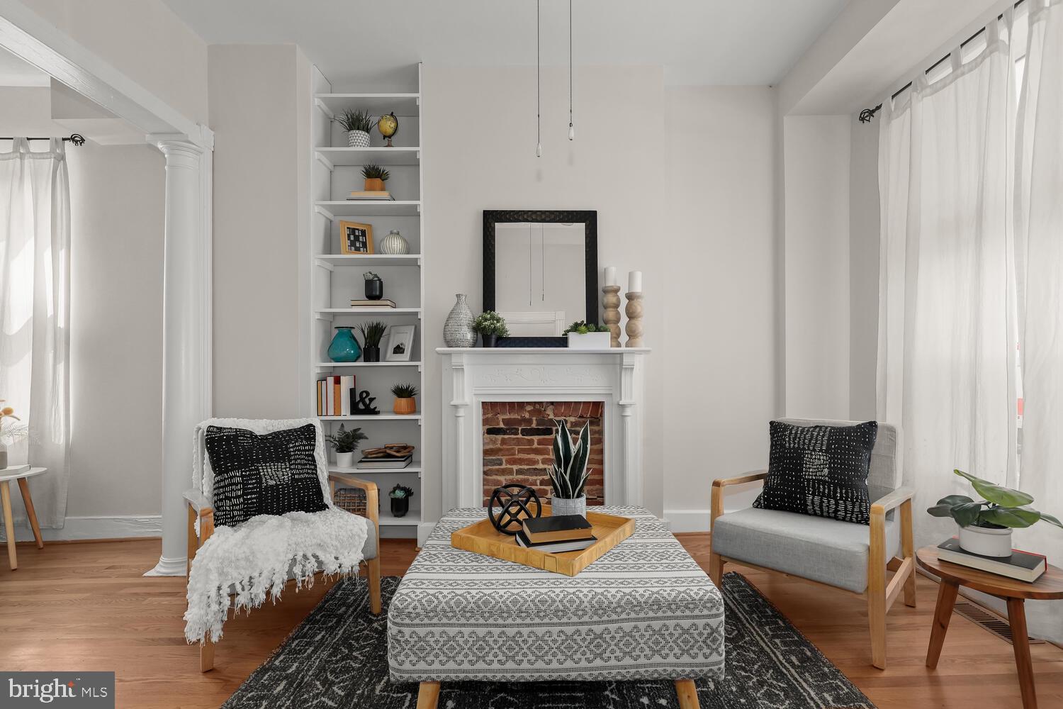Staged living room with a brick fireplace, built-in shelves, and two armchairs around a patterned ottoman.